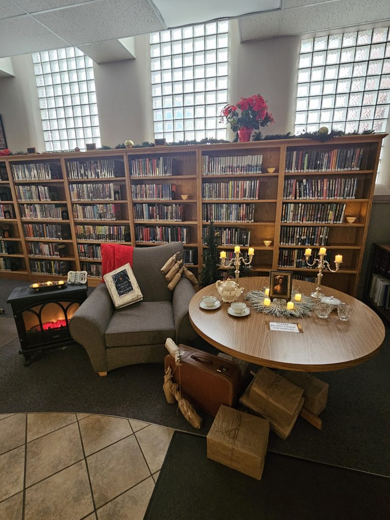 A round table with a tea set, two candelabras, and picture of a faun as the centerpiece surrounded by votive candles. Under the table is a suitcase with brown paper-wrapped milk bottles draped over it next to brown paper-wrapped packages. To the left of the table is an armchair with more milk bottles draped over the back, a red scarf, and a giant Chronicles of Narnia book. To the other side of the chair is an electric fireplace with a decorative plaque and more votive candles on top. In the background are bookshelves with ornaments and garland across the top and a poinsettia plant.