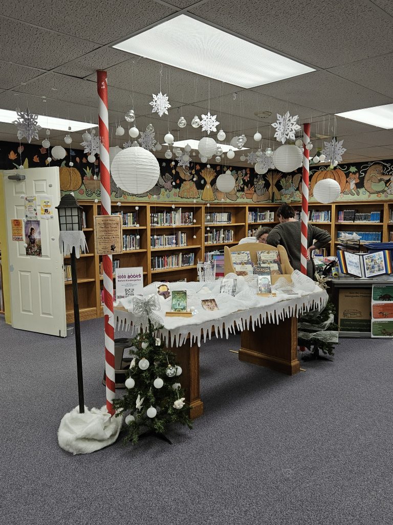 The picture shows a table covered in white fabric and ribbon to depict snow drifts with books on top and felt icicles hanging off the edges. C.S. Lewis Narnia books decorate the snowscape as well as a handmade icicle crown like the Queen's in Narnia. On either side of the table are candy can striped poles in red and white and short Christmas trees. Above the table are a bunch of snowflakes and white baubles to depict falling snow. To the left of the table, on the left pole, is a warrant for Mr. Tumnus's arrest and next to the pole is a lamppost with felt icicles and a felt snow pile at the base. In the background are shelves of books and the youth librarian's desk.